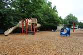 This outdoor playground offers a safe and engaging environment designed for active play. The ground is fully covered with a generous layer of wood chip mulch, providing a natural, impact-absorbing surface that promotes safety throughout the play area. A standout feature is the multi-component play structure positioned on one side of the space, which includes a prominent spiral slide and an attached climbing element, offering children multiple ways to engage and explore. Nearby, a brightly colored blue and yellow seesaw provides an interactive option for group play, adding variety to the recreational equipment on offer. A perimeter of tall, mature trees surrounds the space, creating a verdant natural backdrop that offers shade and a sense of seclusion. The entire area is enclosed by a sturdy metal fence, ensuring a secure environment for unsupervised play. A bench is also visible along the fence line, providing a comfortable seating area for caregivers and observers. The overall ambiance is welcoming and functional, blending natural greenery with purposeful play equipment to create an inviting destination for children of various ages.