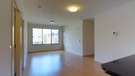 This single-floor space offers a clean, modern aesthetic defined by light hardwood flooring that extends throughout the open living area. The room is bathed in natural light, courtesy of a large multi-pane window that frames views of the surrounding urban neighborhood, including neighboring buildings and mature trees. The white walls and neutral tones create a bright, airy atmosphere that enhances the sense of openness.

Overhead, flush-mount ceiling light fixtures provide consistent ambient illumination, complementing the natural light from the window. The smooth, white ceilings contribute to the room's clean and uncluttered feel. Crisp white baseboards line the perimeter, adding a polished finishing detail throughout.

The open layout offers flexibility for various furniture arrangements, making it well-suited for a living or dining area configuration. Electrical outlets are conveniently positioned along the walls, and a wall-mounted control panel is visible near the window area. The space is move-in ready, presenting a blank canvas with a welcoming, well-maintained character ideal for personalizing to one's taste.
