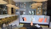 This single-floor space makes a strong visual impression through its double-height interior, where an upper gallery wall displays an eclectic arrangement of framed artwork and photographs, adding character and depth to the environment. The design aesthetic blends contemporary and bohemian influences, anchored by a series of large, woven rattan pendant lights that hang at varying heights, casting a warm, ambient glow throughout the space.

The main seating area features a low-profile sofa adorned with colorful accent cushions in coral and navy tones, paired with a dark coffee table that grounds the arrangement. A terracotta-toned vase with delicate dried botanicals serves as a centerpiece, adding a natural, organic element to the setting.

The walls are finished in a muted blue-grey tone, providing a calm backdrop that complements the warm textures of the lighting and furnishings. To one side, open shelving displays an organized collection of jars or containers, suggesting a functional retail or café component. Large windows along the upper level allow natural light to filter in, enhancing the open, airy atmosphere of this thoughtfully designed space.