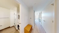 This single-floor space showcases a clean, contemporary design characterized by crisp white walls, light-toned hardwood flooring, and a bright, minimalist aesthetic carried consistently throughout. The open living area benefits from floor-to-ceiling glass doors that lead to a private balcony, flooding the space with natural light and providing open views of the surrounding neighborhood skyline.

The kitchen is well-appointed with flat-front white cabinetry, light quartz countertops, a textured white tile backsplash, an undermount sink with a modern pull-down faucet, a built-in stainless steel oven, an induction cooktop, an integrated microwave, and a stainless steel refrigerator. Open shelving provides additional storage and display space.

The bathroom features a wood-toned vanity with a white quartz countertop, an undermount sink, and mirrored medicine cabinets above. A glass-enclosed tiled shower is accessible nearby.

A dedicated hallway nook accommodates a stacked washer and dryer unit, keeping laundry functions discreetly tucked away. Track lighting and a thermostat panel are noted practical details. The layout, as revealed by the floorplan view, is efficiently organized, connecting the living, kitchen, bathroom, and bedroom areas through a central corridor.