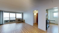 This well-appointed one-bedroom, one-bathroom residence offers a comfortable and efficient layout across a single floor. The living area is bathed in natural light, courtesy of a large sliding glass door that opens to a private balcony, offering views of the surrounding environment. Light-toned hardwood flooring runs throughout the space, lending warmth and continuity to the interior. The clean white walls and flat ceilings create a bright, airy ambiance that makes the space feel open and welcoming. A wall-mounted air conditioning unit ensures year-round comfort within the living area. A hallway leads from the main living space to the bedroom and bathroom, maintaining a clear and practical flow throughout the unit. Warm wall sconce lighting in the hallway adds a subtle accent and enhances the transition between rooms. The overall design is clean and minimal, providing a neutral canvas that adapts easily to a variety of furnishing styles. With its combination of natural light, hardwood flooring, and access to an outdoor balcony, this residence delivers a functional yet comfortable living experience.