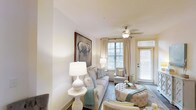 This single-floor residence features a thoughtfully designed living area that combines comfort and style. The living room is finished in a soft, neutral color palette dominated by crisp whites and warm creams, creating a clean and inviting atmosphere. Smooth white walls and a white ceiling contribute to the sense of openness and airiness throughout the space.

Natural light is a defining characteristic of the living room, with multiple windows dressed in light-filtering blinds and sheer curtains allowing sunlight to fill the room. A glass-paneled door provides additional brightness and likely offers access to an outdoor area. A ceiling fan with a light fixture is centrally positioned, adding both functionality and a subtle decorative element.

The living area is furnished with a plush sofa accented with teal and neutral throw pillows, complemented by tufted ottomans and an upholstered accent chair, all arranged around a soft area rug. A large framed artwork featuring an elephant serves as a striking focal point on one wall, adding visual interest. Table lamps with white ceramic bases provide warm ambient lighting, enhancing the overall elegance and cohesion of the space.