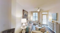 This single-floor residence features a thoughtfully designed living area that combines comfort and style. The living room is finished in a soft, neutral color palette dominated by crisp whites and warm creams, creating a clean and inviting atmosphere. Smooth white walls and a white ceiling contribute to the sense of openness and airiness throughout the space.

Natural light is a defining characteristic of the living room, with multiple windows dressed in light-filtering blinds and sheer curtains allowing sunlight to fill the room. A glass-paneled door provides additional brightness and likely offers access to an outdoor area. A ceiling fan with a light fixture is centrally positioned, adding both functionality and a subtle decorative element.

The living area is furnished with a plush sofa accented with teal and neutral throw pillows, complemented by tufted ottomans and an upholstered accent chair, all arranged around a soft area rug. A large framed artwork featuring an elephant serves as a striking focal point on one wall, adding visual interest. Table lamps with white ceramic bases provide warm ambient lighting, enhancing the overall elegance and cohesion of the space.