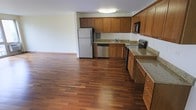 This single-floor space is defined by an open, airy layout that seamlessly connects the kitchen to the adjoining living area, creating a generous and functional flow. The kitchen is well-appointed with warm honey-toned wood cabinetry that extends to the upper walls, providing ample storage. Granite countertops in a light, speckled finish add a durable and attractive work surface, complementing the cabinetry's natural tones. A full suite of stainless steel appliances includes a refrigerator, electric range, built-in microwave, and dishwasher, offering modern convenience throughout. The sink is centrally positioned beneath upper cabinets, making the workspace efficient and practical. Rich, dark hardwood flooring spans the entire visible area, lending warmth and a cohesive aesthetic to the space. The open layout beyond the kitchen provides a spacious living zone, illuminated by flush-mount ceiling lighting. White walls keep the interior feeling bright and neutral, allowing the natural wood tones of both the cabinetry and flooring to serve as the primary design elements. Overall, the space presents a clean, functional, and inviting environment.