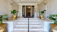 The Claremont House is a distinguished building defined by its timeless classical architecture and refined interior detailing. The grand entrance is approached via a sweeping marble staircase flanked by geometric gold planters with lush tropical foliage, setting a stately tone from the moment of arrival. Black-framed glass doors with ornate ironwork open into a vestibule clad in veined marble walls, providing a refined transition into the main lobby.

The lobby itself is a showpiece of architectural craftsmanship, featuring an elaborately detailed plaster ceiling medallion, deep decorative crown moldings, and a central chandelier with warm amber globe shades. Marble-clad walls continue throughout, complemented by sleek benches and potted greenery that add a welcoming touch. Tall glass entry doors frame views of the surrounding streetscape, filling the space with natural light.

Beyond the lobby, a wide carpeted corridor extends through the building, lined with paneled teal doors and accented by ornate ceiling cornices that echo the classical detailing found at the entrance. A staircase with dark-painted wooden balustrades and light wood treads provides access to upper levels, positioned at the corridor's midpoint. The overall ambiance is one of historic elegance, combining original architectural elements with a well-maintained, functional interior.