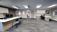 This single-floor office space presents a clean, contemporary design well-suited for professional use. The main area functions as an open reception and workstation zone, finished with neutral taupe walls and patterned grey carpet tiles that lend a polished, business-ready atmosphere. Flush-mounted ceiling light panels provide consistent, even illumination throughout the space.

A curved reception desk with guest seating occupies one side of the room, complemented by a built-in service window with shelving — ideal for client-facing interactions. On the opposite side, a dedicated workstation area features built-in upper cabinetry in white, a two-tone desk with drawer storage, and space for computer equipment, supporting day-to-day administrative tasks.

A doorway visible from the main area leads to an additional room, which appears to offer supplemental workspace or meeting space with its own seating and wall art. The layout is open and uncluttered, promoting ease of movement and a welcoming professional environment. Overall, the space balances functionality with a modern aesthetic throughout.