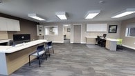 This single-floor office space presents a clean, contemporary design well-suited for professional use. The main area functions as an open reception and workstation zone, finished with neutral taupe walls and patterned grey carpet tiles that lend a polished, business-ready atmosphere. Flush-mounted ceiling light panels provide consistent, even illumination throughout the space.

A curved reception desk with guest seating occupies one side of the room, complemented by a built-in service window with shelving — ideal for client-facing interactions. On the opposite side, a dedicated workstation area features built-in upper cabinetry in white, a two-tone desk with drawer storage, and space for computer equipment, supporting day-to-day administrative tasks.

A doorway visible from the main area leads to an additional room, which appears to offer supplemental workspace or meeting space with its own seating and wall art. The layout is open and uncluttered, promoting ease of movement and a welcoming professional environment. Overall, the space balances functionality with a modern aesthetic throughout.