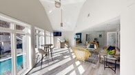 Located at 3131 Mountain Creek Rd in Chattanooga, Tennessee, this single-floor living space offers a bright, open-concept design that maximizes both comfort and visual appeal. The centerpiece of the room is a dramatic vaulted ceiling that rises to an impressive peak, creating a grand sense of openness and volume. A modern ceiling fan anchors the apex, complementing the clean, white-painted walls that reflect natural light throughout the day.

An expansive wall of floor-to-ceiling windows lines one side of the space, flooding the interior with abundant sunlight and offering direct views of an outdoor pool area and the surrounding landscape — a standout architectural feature that seamlessly connects the indoors with the outdoors. Light wood-toned luxury vinyl flooring runs throughout, adding warmth and continuity to the space.

The living area is thoughtfully arranged with distinct zones. A casual lounge area features woven rattan chairs in bold yellow, a glass-top coffee table, and a sofa set against a striking teal accent wall adorned with contemporary artwork. A wall-mounted flat-screen TV and a herringbone-patterned wood credenza provide both functional and decorative interest. A bistro-height table with bar stools near the windows creates an ideal spot for casual seating with pool views. Large mirrored panels further amplify the sense of space and light throughout.