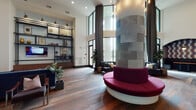 This thoughtfully designed single-floor amenity space combines sophisticated aesthetics with functional comfort throughout its distinct zones. The grand lobby lounge immediately impresses with soaring double-height ceilings, rich hardwood flooring, and an eye-catching cylindrical stone column wrapped in stacked gray tile. A signature circular crimson bench wraps around the column, serving as a bold focal point. Floor-to-ceiling windows dressed with dramatic floor-length drapes flood the space with natural light while framing lush outdoor views of a landscaped courtyard with palm trees. Industrial-style open shelving with a rolling library ladder adds a curated, editorial touch alongside plush seating arrangements.

Adjacent to the lounge, a fully equipped kitchen and social bar area showcases teal cabinetry with decorative lattice upper cabinets, marble-look countertops, and a sleek bar with black bistro-style stools. Intimate arched booth alcoves with patterned upholstery and moody pendant lighting provide semi-private seating, ideal for small gatherings or relaxed dining.

A compact business nook features a wall-mounted white desk, task seating, and a printer station set against a calming light blue wall. Throughout, warm hardwood floors, high ceilings, and a cohesive modern-eclectic palette create an inviting and versatile communal environment.