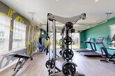 Located at 1702 Rocky River Road in Charlotte, NC, this fitness center is a thoughtfully designed exercise space that delivers a comprehensive workout environment across a single floor. The interior is finished with durable wood-look flooring and vibrant teal accent walls, complemented by an eye-catching motivational mural featuring bold yellow typography that energizes the space. Recessed lighting throughout ensures clear visibility across all workout zones.

The cardio area is equipped with multiple treadmills, an elliptical, and a stationary bike, all positioned alongside windows that bring in natural light. A dedicated strength training zone features a full cable and functional trainer system loaded with weight plates, along with a TRX suspension trainer for bodyweight exercises. Free weights, including a full dumbbell rack, adjustable benches, and plyometric boxes, round out the strength offerings. Stability balls, resistance accessories, and wall-mounted exercise guides support functional and flexibility training.

A wall-mounted flat-screen TV and a water fountain add convenience for users. One of the standout architectural features is the large garage-style door that opens fully to an adjacent outdoor turf area, seamlessly extending the workout space and flooding the interior with fresh air and natural light. The exterior grounds are neatly landscaped with manicured turf, ornamental shrubs, and iron fencing, creating an inviting and polished environment.