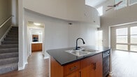 Located at 1101 Bear Creek Pkwy in Keller, Texas, this two-story residence showcases a distinctive architectural design highlighted by dramatic curved walls and soaring double-height ceilings that define the main living area. Rich hardwood flooring flows throughout the first floor, complementing the open-concept layout that seamlessly connects the living and kitchen spaces. The kitchen features a central island with dark granite countertops, a double stainless steel sink with a matte black faucet, warm wood cabinetry, and black appliances including a side-by-side refrigerator. Large windows and a sliding glass door with vertical blinds invite abundant natural light into the living area, enhancing the sense of openness. A staircase leads to the second floor, where carpet flooring adds comfort to the upper-level rooms. The upper floor includes a spacious loft-style area with a ceiling fan, vaulted ceiling, and multiple windows, as well as a bedroom with a closet and access to additional rooms. A bathroom on the lower level is outfitted with tile walls, a glass-enclosed walk-in shower, wood-toned cabinetry, and hardwood flooring. Throughout both floors, neutral tones and clean lines create a cohesive, modern aesthetic.