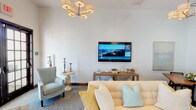 This single-floor communal lounge space exudes a refined yet comfortable ambiance, blending classic architectural elements with contemporary coastal-inspired décor. Upon entry, grand dark-wood double doors with transom windows usher guests into a generously proportioned open area bathed in natural light. Soaring ceilings amplify the sense of openness, while a multi-arm chandelier with drum shades adds a polished focal point overhead.

The main lounge area is anchored by a dramatic floor-to-ceiling stone fireplace with a rustic wooden mantle, lending warmth and texture to the otherwise crisp, white-walled interior. Comfortable seating — including a cream tufted sofa dressed with soft aqua and yellow accent pillows and neutral wingback chairs — creates a welcoming gathering environment. A wall-mounted flat-screen television and a wood-and-metal media console complete the entertainment corner.

Black-framed French doors connect the main lounge to additional seating areas and adjacent rooms, maintaining visual flow while defining distinct zones. Throughout the space, plush area rugs, accent tables, and carefully placed décor pieces contribute to the cohesive, coastal aesthetic. Natural light filters in generously through multiple sets of French doors, offering views to an outdoor area beyond.
