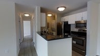 This single-floor space centers around a well-appointed kitchen that combines functionality with a clean, modern aesthetic. The kitchen features a peninsula with a dark, reflective countertop that provides additional prep space and serves as a natural divider between the cooking area and the rest of the living space. White shaker-style cabinetry lines the upper walls, offering ample storage, while stainless steel appliances — including a refrigerator, microwave, and range — add a contemporary touch. A single-handle arc faucet is integrated into the peninsula, adding both style and convenience.

The flooring throughout is a wood-look material in a neutral tone, contributing to a cohesive and easy-to-maintain interior. Flush-mount ceiling light fixtures provide consistent overhead lighting across the kitchen and connecting hallway. A stacked washer and dryer unit is visible in a dedicated laundry nook adjacent to the kitchen, maximizing the use of available space. The hallway extending from the kitchen leads to additional rooms, all presented with clean white walls and consistent flooring, giving the overall space a bright, move-in-ready feel.