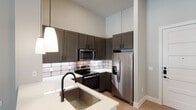 This single-floor unit showcases a thoughtfully designed interior with a contemporary aesthetic and high-end finishes throughout. The soaring ceilings create an expansive, airy atmosphere that is immediately noticeable upon entry, amplified further by the abundant natural light streaming through large windows fitted with white horizontal blinds.

The kitchen is a standout feature, equipped with rich, dark taupe shaker-style cabinetry extending to the upper walls to maximize storage. A classic white subway tile backsplash adds a clean, timeless contrast, while the white quartz countertops provide ample prep space. Stainless steel appliances — including a French door refrigerator with ice maker, an electric range, a built-in microwave, and a dishwasher — complete the highly functional cooking space. Pendant lighting hangs elegantly over the kitchen peninsula, which features a deep undermount sink with a matte black faucet.

The open layout connects the kitchen seamlessly to the living area, where warm wood-look flooring and a ceiling fan contribute to a comfortable, casual ambiance. The bedroom area offers carpeted flooring and an outdoor view through a large window. The bathroom includes a tiled soaking tub, wood-look tile flooring, and a well-organized walk-in closet with double hanging rods, offering generous storage for everyday living.