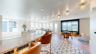 Located in Orlando, FL, this thoughtfully designed communal space offers a sophisticated and welcoming atmosphere across a single, well-appointed floor. The interior showcases a contemporary aesthetic defined by clean lines, warm wood flooring, and an eye-catching geometric patterned tile that anchors the main gathering areas.

At the heart of the space is a sweeping curved bar island clad in teal green tile with a granite countertop and brass-accented globe pendant lighting overhead. Leather barrel-back bar stools invite casual seating, while an adjacent lounge area features plush, rounded armchairs arranged around a central coffee table beneath statement brass drum pendant lights. A slatted wood accent wall frames a large wall-mounted television, while a bold teal zellige tile feature wall serves as a striking gallery display backdrop.

Large windows along multiple walls flood the interior with abundant natural light and offer open views of the surrounding neighborhood. A dedicated seating nook near the windows provides additional intimate gathering space with bistro-style tables and chairs.

The space extends to a private balcony where residents can enjoy the Florida sunshine alongside sweeping views of the community's modern residential buildings. A closet rounds out the layout, adding practical functionality to this beautifully curated space.