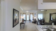 This single-floor residence offers an open, flowing layout that connects the kitchen, dining, and living areas in a cohesive and functional arrangement. The interior is finished with elegant crown molding along the ceilings, contributing a classic, refined character throughout the space. Neutral wall tones and light-colored carpeting in the living area create a warm and inviting ambiance.

The living room serves as the focal point, featuring a seating arrangement centered around a chaise lounge and accent chairs. Natural light pours in generously through multiple windows dressed with gold-toned drapes, as well as a full glass exterior door that leads to an outdoor area, enhancing the sense of openness. The large framed artwork displayed along the walls adds a decorative touch and visual interest.

Adjacent to the living space, the dining area accommodates a dining table with upholstered chairs, positioned near additional windows that further brighten the area. The kitchen is partially visible, featuring light countertops and a sink, seamlessly integrated into the overall open-concept design. Recessed and flush-mount lighting supplement the natural light, ensuring the space remains well-lit throughout.