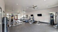 Located at 3815 NE 4th St in Renton, WA, this well-equipped fitness center offers a spacious, single-floor layout designed to support a full range of workout routines. The interior features durable rubber flooring throughout, providing a safe and functional surface for exercise. The space is organized to accommodate both cardio and strength training, with multiple treadmills, elliptical machines, stationary bikes, and a rowing machine positioned alongside a cable weight machine, free weights, and adjustable benches. Several wall-mounted flat-screen TVs are strategically placed to keep users entertained during workouts. Overhead, multiple modern ceiling fans complement the recessed lighting, ensuring consistent airflow and a comfortable environment. The color palette features crisp white walls contrasted by a bold dark accent wall, lending the space a contemporary aesthetic. Large windows and a set of double glass entry doors flood the room with natural light while offering views of the surrounding outdoor area. The facility's clean, open layout maximizes usable floor space, making it easy to navigate between equipment. Overall, this fitness center provides a functional, modern, and well-lit environment suited to a variety of fitness needs.