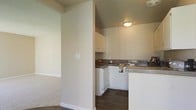 This single-floor residence offers a practical and well-organized layout. The kitchen is a standout feature, designed with white cabinetry that provides ample storage space, including upper wall-mounted cabinets. Dark-toned countertops contrast neatly with the white surfaces, while a built-in dishwasher and sink contribute to the kitchen's functional appeal. The flooring in the kitchen area is dark hardwood or wood-look material, adding warmth and a modern touch. A flush-mount ceiling light provides focused illumination throughout the space. Adjacent to the kitchen, a partial wall separates the cooking area from the neighboring room, which features soft wall-to-wall carpeting, creating a comfortable and distinct living area. The neutral color palette of warm beige and off-white tones throughout the space gives the interior a cohesive, understated aesthetic. The layout encourages an easy transition between the kitchen and the adjoining carpeted area, making the space feel connected while maintaining defined zones for different activities. Overall, the residence presents a clean, move-in-ready environment with practical finishes suited for everyday living.