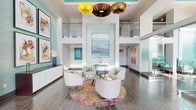 This contemporary space makes an immediate impression with its dramatic double-height ceilings and open, airy design. The main living area is anchored by a striking cluster of sculptural pendant lights — featuring yellow, brown, and bronze tones — that hang as artistic focal points above a curated seating arrangement. Four sleek white swivel chairs surround a glass coffee table, all set atop a vivid, multicolored circular rug that adds a bold splash of warmth and energy to the refined space.

The walls are adorned with carefully selected artwork, complementing the soft aqua and teal accent tones that appear throughout, including a shimmering mosaic tile feature wall framing a large abstract painting. Clean white walls and wide-plank hardwood-style flooring provide a neutral backdrop that allows the colorful design elements to truly stand out.

A glass-railed upper-level overlook adds architectural depth and visual interest, enhancing the sense of vertical space. Natural light flows generously throughout, further brightening the interior. A sleek white credenza along one wall provides functional storage while maintaining the space's polished aesthetic. Overall, the space exudes a modern, gallery-like ambiance with a sophisticated yet approachable character.
