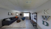 This single-floor residence offers a comfortable and thoughtfully arranged living space. The living room serves as the heart of the home, featuring light wood-style flooring that extends throughout and a textured ceiling that adds a classic touch. A large, plush area rug anchors the seating area, which includes a spacious dark gray sofa accented with bold orange throw pillows and a complementary blue accent chair. A rustic wood coffee table ties the seating arrangement together, while a floor lamp provides ambient lighting. A window dressed with sheer white curtains allows natural light to filter into the room, creating a bright and airy atmosphere. A decorative wall shelf with a small piece of artwork adds a personal touch to the space. Near the entryway, a sleek industrial-style coat rack and bench unit offers practical storage for outerwear and accessories, making for a clean and organized arrival area. The dark front door provides a defined entry point, while the HVAC vent integrated into the ceiling ensures comfortable climate control throughout. Overall, the space balances functional design with a warm, inviting ambiance suited for everyday living.