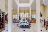 Located at 150 Du Rhu Dr in Mobile, Alabama, this distinguished single-floor space makes an immediate impression with its grand, column-framed foyer. Soaring ceilings—accented by an upper clerestory window arrangement—flood the interior with natural light, while a classic chandelier anchors the central gathering area. Light-toned tile flooring with decorative inlays runs throughout, enhancing the sense of openness and flow.

The main living area is generously proportioned, featuring warm yellow walls, a fireplace with a white mantel, and abundant French doors that frame lush outdoor views and invite natural light from multiple directions. Comfortable, traditional furnishings complement the refined architectural details including crown molding and tall white columns.

A well-appointed kitchenette offers dark granite countertops, medium-toned wood cabinetry, a sink, and bar-height seating—ideal for casual gatherings or serving guests. Adjacent hallways display framed artwork and connect seamlessly to additional rooms throughout the space.

The covered outdoor area extends the living experience further, featuring a fully equipped outdoor kitchen with a large grill and commercial-style range hood, ceiling fans, a mounted television, and multiple dining sets overlooking a manicured landscape. Together, these spaces combine elegant interior design with practical amenities in a welcoming, polished environment.