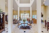 Located at 150 Du Rhu Dr in Mobile, Alabama, this distinguished single-floor space makes an immediate impression with its grand, column-framed foyer. Soaring ceilings—accented by an upper clerestory window arrangement—flood the interior with natural light, while a classic chandelier anchors the central gathering area. Light-toned tile flooring with decorative inlays runs throughout, enhancing the sense of openness and flow.

The main living area is generously proportioned, featuring warm yellow walls, a fireplace with a white mantel, and abundant French doors that frame lush outdoor views and invite natural light from multiple directions. Comfortable, traditional furnishings complement the refined architectural details including crown molding and tall white columns.

A well-appointed kitchenette offers dark granite countertops, medium-toned wood cabinetry, a sink, and bar-height seating—ideal for casual gatherings or serving guests. Adjacent hallways display framed artwork and connect seamlessly to additional rooms throughout the space.

The covered outdoor area extends the living experience further, featuring a fully equipped outdoor kitchen with a large grill and commercial-style range hood, ceiling fans, a mounted television, and multiple dining sets overlooking a manicured landscape. Together, these spaces combine elegant interior design with practical amenities in a welcoming, polished environment.