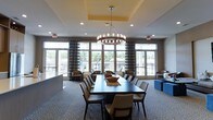 This single-floor communal space is designed to accommodate group gatherings and social events in a refined, comfortable setting. The room is anchored by a generously sized dark wood dining table surrounded by upholstered chairs, making it well-suited for formal dinners or meetings. Overhead, a striking chandelier with candle-style bulbs serves as a focal point, suspended from a tray ceiling accented with warm yellow paint that adds a subtle pop of color to the neutral palette.

The space benefits greatly from an expansive wall of floor-to-ceiling windows and glass doors, allowing abundant natural light to flood the interior and offering clear views of an outdoor terrace or patio area beyond. Plaid-patterned drapes frame the windows, adding a classic, tailored touch. The flooring features a patterned carpet in soft, muted tones that complements the overall neutral color scheme.

Additional seating, including armchairs and a blue upholstered ottoman, is arranged near the windows, creating a relaxed lounge area alongside the formal dining setup. Recessed lighting throughout the ceiling supplements the natural light, ensuring the space remains bright and inviting at all times.