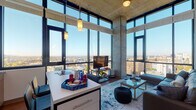 This contemporary high-rise residence offers four bedrooms and two bathrooms across a single thoughtfully designed floor. The open-concept living and kitchen area immediately impresses with soaring floor-to-ceiling windows that wrap around the space, flooding the interior with natural light and framing expansive panoramic city views. Exposed concrete columns and ceilings add an industrial character that contrasts beautifully with the clean white finishes and warm wood-look flooring found throughout.

The kitchen is well-appointed with two-tone cabinetry — crisp white uppers paired with natural wood lowers — a subway tile backsplash, stainless steel appliances including a dishwasher and microwave, quartz-style countertops, and a central island with seating for casual dining. Pendant lighting accents both the kitchen and living areas.

The living room features a large sectional sofa, stylish area rug, and curated wall art, all set against a backdrop of sweeping skyline views. Each bedroom benefits from the building's generous window line, offering bright, airy spaces with city vistas. The bathrooms are finished with wood-toned vanities and neutral countertops. A clean, well-lit hallway connects the private quarters, complete with decorative shelving and a mirror. This residence combines modern urban design with comfortable, functional living.