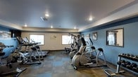 This fitness center offers a thoughtfully arranged workout space designed to accommodate a range of exercise needs. The room features a combination of cardio machines, including treadmills and ellipticals, as well as a multi-function cable/weight machine, providing users with both cardiovascular and strength-training options in a single space.

The interior is finished with a two-tone color scheme — warm beige on the upper walls transitioning to a cool steel blue on the lower section — giving the room a clean, energizing aesthetic. Recessed lighting throughout the ceiling ensures consistent, well-distributed illumination across the workout area. The flooring consists of durable, multi-toned carpet tiles that provide both comfort and practicality underfoot.

Natural light filters in through several windows along one wall, contributing to an open and inviting atmosphere. Motivational artwork and framed prints are displayed along the blue accent wall, adding character to the space. A vinyl wall decal reads an inspirational quote, reinforcing the fitness-focused environment. The single-floor layout ensures easy accessibility throughout.