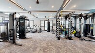 Located at 50 Fenway Place in Concord, NC, this well-appointed fitness center is designed to support a full range of workout disciplines within a thoughtfully organized, single-floor layout. The main strength and cardio area features textured carpet flooring, coffered ceilings with recessed lighting and accent trim, and ceiling fans that promote air circulation throughout. This zone is equipped with a multi-function cable machine, a power rack loaded with barbells and weight plates, a full dumbbell rack, a heavy punching bag, treadmills including an Assault Fitness model, and an upright stationary bike. A dedicated cycling and studio room offers light wood-look tile flooring, large windows with views of an outdoor pool area, a floor-to-ceiling mirror wall with a large mounted display screen, spin bikes, stability balls, resistance ropes, and a step platform — ideal for group fitness or individual training sessions. Natural light flows generously through multiple windows fitted with adjustable blinds throughout both zones. Near the entrance, a wooden cubby storage unit and a designated Fitness Center sign provide an organized welcome point. The overall aesthetic is clean and modern, with a neutral gray and blue color palette that creates an energizing yet focused environment for exercise.