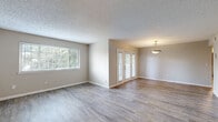 This single-story home presents a clean, neutral aesthetic with consistent design choices throughout. The open living and dining area is finished with light wood-look luxury vinyl flooring and features a large window that draws in natural light with views of surrounding greenery. French doors connect the dining area to the exterior, enhancing the sense of openness, while a pendant light fixture adds a subtle decorative touch. A textured ceiling is present in the main living areas.

A hallway with wood-look flooring leads to the private areas of the home, where multiple bedrooms are finished with plush carpet. Each bedroom includes a window overlooking lush outdoor greenery, and most are equipped with ceiling fan and light combinations. One bedroom features a built-in closet with sliding doors. The neutral wall tones create a calm, versatile backdrop throughout.

The bathroom includes wood-look tile flooring, a single-sink vanity with warm wood cabinetry and a dark granite-style countertop, a medicine cabinet mirror, and a tiled tub-shower combination. The hallway connecting the rooms is well-lit and features a textured ceiling, maintaining the home's cohesive, understated style.