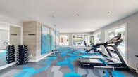 Located at 14801 Lemoyne Blvd in Biloxi, Mississippi, this fitness center offers a thoughtfully designed workout environment within a single open floor. The space is defined by its eye-catching geometric carpet tiles in bold shades of blue and gray, which give the room a modern, energetic aesthetic that complements the coastal surroundings.

Large picture windows line the walls, flooding the interior with natural light and providing clear views of the outdoor pool area and surrounding grounds. A wall-mounted flat-screen TV and recessed ceiling lighting add to the functional, well-lit atmosphere.

The exercise room is comprehensively equipped for a full range of workouts. Cardio equipment includes treadmills, a rowing machine, and a stationary spin bike. A dedicated free weights area features a full dumbbell rack alongside a weight bench, while a multi-station cable machine supports strength and resistance training. Stability and balance tools, including medicine balls and a balance disc, round out the functional fitness offerings.

A striking large-format triptych mural of an ocean wave serves as a bold focal point, reinforcing the space's coastal character. A hallway corridor with additional natural light leads through the facility, accented by decorative greenery. The space is accessible via glass-paned doors that open to the outdoor amenity area, seamlessly connecting indoor fitness with the outdoor environment.