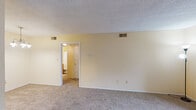 This single-floor residence offers a practical and comfortable layout, finished throughout in warm yellow and cream tones that lend a welcoming ambiance to every room. The entryway opens into a spacious living area with plush wall-to-wall carpeting, a sliding glass door that invites natural light and outdoor access, and ample wall space for flexible furniture arrangement. Adjacent to the living area, the kitchen is equipped with warm oak cabinetry, a white electric coil range with overhead ventilation, a full-size refrigerator, and dark laminate countertops. The durable wood-look vinyl flooring in the kitchen adds contrast and easy maintenance. A bedroom features carpet flooring, a ceiling fan with integrated light fixture, a window with blinds for natural light and ventilation, and a connected hallway leading to the bathroom. The bathroom includes a single-sink vanity with a white basin, oak cabinetry below, a large mirror with overhead lighting, and textured wall finishes. A separate laundry area with washer and dryer hookups adds functional convenience. Textured ceilings are present throughout, and central HVAC vents are consistently placed for climate control across the space.