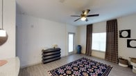 This single-floor space offers a clean, contemporary layout designed for comfortable everyday living. The main room is finished with light wood-style flooring that runs throughout, lending a warm and cohesive feel to the interior. A five-blade ceiling fan with an integrated light fixture is centrally mounted, providing both ambient lighting and efficient air circulation year-round.

Natural light is a defining feature of this space, with large windows dressed in patterned curtains that allow for both brightness and privacy. A glass-paneled door provides additional light and direct access to an exterior area, enhancing the sense of openness. The neutral white walls serve as a versatile backdrop, complementing a range of furnishing styles.

A bold, patterned area rug in deep navy with floral motifs adds a vibrant focal point to the room, while a dark-finished multi-drawer dresser contributes functional storage without compromising the aesthetic. A decorative blue ceramic vase adds a touch of artful character near the window. The overall ambiance is bright, airy, and welcoming, with a thoughtful balance of practicality and style that makes the space well-suited for a variety of uses.