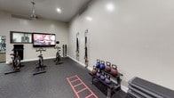 This dedicated CrossFit and functional fitness space is thoughtfully designed to support a wide range of training modalities in a single, open-plan room. The interior features durable rubber flooring throughout, providing the safety and grip necessary for high-intensity workouts, complemented by clean white walls and recessed lighting alongside a stylish pendant ceiling fan that keeps the space well-lit and ventilated.

A full-length wall mirror spans a significant portion of one side of the room, enhancing the sense of openness and allowing users to monitor their form during exercise. A wall-mounted flat-screen TV is positioned for easy viewing from multiple workout zones. The space is outfitted with an impressive selection of equipment including spin bikes, a multi-functional cable and suspension training rig, TRX straps, battle ropes, kettlebells of varying weights, medicine balls, stability balls, a BOSU balance trainer, resistance bands, a speed ladder, step platforms, and a dumbbell rack. Wall-mounted storage keeps equipment organized and accessible. The high ceilings contribute to an open, airy atmosphere well-suited for dynamic movements and group training sessions, making this a comprehensive and professional-grade fitness environment.