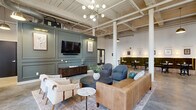 Located in Augusta, Georgia, this clubhouse masterfully combines industrial heritage with contemporary design. The single-floor space features soaring ceilings with exposed structural beams, painted white brick walls, and polished concrete floors that run throughout, creating a cohesive and airy atmosphere.

The lounge area anchors the space with a sage-green paneled accent wall housing a mounted flat-screen TV, flanked by framed botanical artwork. Comfortable seating — including a cognac leather sofa, upholstered armchairs, and a marble-top accent table — is arranged around a dark round coffee table atop a layered area rug. A striking mid-century modern chandelier with globe bulbs illuminates the area warmly.

Adjacent to the lounge, a dining nook features tufted green velvet banquette seating paired with round marble-top tables and black cross-back chairs, accented by brass wall sconces and curated wall art. Tall, arched multi-pane windows flood a separate game and social area with abundant natural light, highlighting whitewashed brick walls and high ceilings. This zone offers communal tables and bar-height seating, ideal for casual gatherings.

A wide-open corridor connects all areas fluidly, reinforcing the expansive, loft-like character of the space. The overall ambiance balances industrial rawness with warm, thoughtfully selected furnishings and lighting.