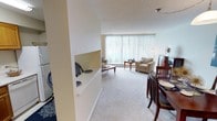 This single-floor residence offers a thoughtfully arranged open-concept layout that seamlessly connects the living, dining, and kitchen areas. The living room is a bright, welcoming space anchored by a large floor-to-ceiling window dressed with vertical blinds, allowing abundant natural light to fill the room while offering pleasant views of the surrounding greenery. Plush wall-to-wall carpeting runs throughout the main living areas, adding warmth and comfort underfoot. A partial wall with a built-in shelf serves as a subtle divider between the kitchen and the living space, providing both visual separation and practical surface area. The dining area accommodates a classic dark wood dining set, positioned to take advantage of the open flow of the room. The textured ceiling adds a layer of architectural detail consistent throughout the space. The kitchen, visible from the entryway, is efficiently arranged and connected to the main living area. Overall, the space presents a clean, neutral aesthetic with light-toned walls and furnishings that enhance the sense of openness. The layout is well-suited for those seeking a comfortable, functional living environment with a cohesive and inviting atmosphere.