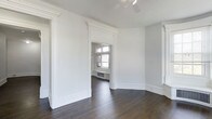 This single-floor space showcases a refined blend of classic architectural character and clean, modern presentation. The interiors are defined by crisp white walls and trim work, with elegantly detailed door surrounds featuring decorative crown molding and pilaster-style casings that speak to the craftsmanship of traditional design. Dark hardwood floors run throughout, providing a rich contrast to the light-toned walls and ceilings, and lending a sense of warmth and depth to each room. The layout flows openly between spaces, connected through wide, architecturally framed doorways that create a strong sense of continuity. Natural light filters in through multi-pane windows visible in the adjoining room, brightening the interior and highlighting the clean lines of the trim and baseboards. The ceilings are notably tall, enhancing the sense of spaciousness and volume throughout. A ceiling-mounted track lighting fixture adds a contemporary functional touch without detracting from the space's classic character. Wide, substantial baseboards complement the overall trim profile and complete the cohesive aesthetic. The overall ambiance is bright, airy, and well-appointed, with architectural details that give the space a distinguished and timeless quality.