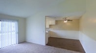This single-floor residence offers a practical and open layout designed for comfortable everyday living. The living area features neutral-toned carpet underfoot and smooth, light-painted walls that create a clean and airy atmosphere. A wide open passageway connects the living space to an adjoining room, which transitions to dark hardwood-style flooring, adding a warm contrast to the overall palette. The kitchen is visible through the open connection and is outfitted with white upper cabinetry, offering a classic and functional design. A built-in counter or pass-through element further integrates the kitchen with the adjacent space, promoting an open and connected feel throughout the home. The adjoining room is equipped with a ceiling fan, providing both practical ventilation and a subtle decorative touch. Textured ceilings run throughout the home, adding dimension to the interior. The neutral color scheme of warm beige and soft white tones creates a cohesive and inviting environment. The layout is thoughtfully designed to maximize flow between living spaces, making the home feel welcoming and versatile for a variety of living arrangements.