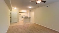 This single-floor space showcases an open-concept layout that seamlessly connects the living area to the kitchen. The living area is finished with neutral-toned carpet, creating a comfortable and inviting atmosphere, while a ceiling fan with an integrated light fixture serves as a functional overhead focal point.

The kitchen occupies the back portion of the space and features white cabinetry providing ample storage, white appliances including a refrigerator, microwave, and range, and a peninsula counter with a darker countertop that acts as a natural dividing element between the two areas. A pendant light hangs above the peninsula, adding a subtle design detail, while recessed-style overhead lighting illuminates the kitchen workspace. The kitchen floor transitions to tile, offering a practical, easy-to-clean surface.

The overall color palette is light and neutral throughout, with soft cream-toned walls and white trim contributing to a bright, clean aesthetic. A paneled door near the kitchen provides access to an additional area of the unit. The open flow between spaces and consistent lighting make this a functional and well-organized layout.