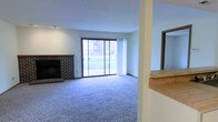 This single-floor space centers around a welcoming living area defined by a classic full-brick fireplace with a raised hearth and a simple wood mantel, serving as a natural focal point. The fireplace is flanked by neutral-toned walls and a textured ceiling, contributing to a clean and understated aesthetic. Plush wall-to-wall carpeting in a muted blue-gray tone covers the living area floor, adding warmth and comfort underfoot. A large sliding glass door dominates one wall, allowing abundant natural light to fill the room while offering direct views of a grassy outdoor area and adjacent greenery. Vertical blinds alongside the sliding door provide adjustable light control and privacy. A partial wall with a wood-toned countertop is visible at the room's edge, suggesting a pass-through or transitional space leading to an adjacent area, with a mirror adding depth to the space. Recessed ceiling fixtures provide supplemental overhead lighting. The overall ambiance is functional and livable, with a straightforward layout that balances natural light, comfortable flooring, and a traditional fireplace feature.