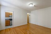 This single-floor unit is thoughtfully laid out, offering a comfortable and functional living environment. The main living area is finished with warm, honey-toned hardwood floors that extend throughout much of the space, complemented by clean white walls and flush ceiling lighting. Baseboard heating runs along the perimeter, and a window-mounted air conditioning unit provides climate control. The open feel of the living space allows for flexible furniture arrangements.

The kitchen is positioned off the main living area and is equipped with solid oak upper and lower cabinetry offering ample storage. Dark granite-style countertops provide a functional work surface, and the kitchen features a stainless steel gas range with an overhead exhaust hood and a single-basin stainless steel sink. A window with blinds brings in natural light, and the floor is finished with a classic black-and-white geometric tile pattern that adds a distinctive visual element.

The bedroom is a well-proportioned, quiet space with the same hardwood flooring found throughout, white walls, and ceiling lighting. The bathroom is finished in a light, neutral palette with tile flooring, a vanity with under-sink storage, a medicine cabinet with mirror, and a toilet. A window allows for natural ventilation. Overall, the unit offers a clean and practical layout suited for everyday living.
