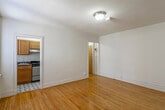 This single-floor unit is thoughtfully laid out, offering a comfortable and functional living environment. The main living area is finished with warm, honey-toned hardwood floors that extend throughout much of the space, complemented by clean white walls and flush ceiling lighting. Baseboard heating runs along the perimeter, and a window-mounted air conditioning unit provides climate control. The open feel of the living space allows for flexible furniture arrangements.

The kitchen is positioned off the main living area and is equipped with solid oak upper and lower cabinetry offering ample storage. Dark granite-style countertops provide a functional work surface, and the kitchen features a stainless steel gas range with an overhead exhaust hood and a single-basin stainless steel sink. A window with blinds brings in natural light, and the floor is finished with a classic black-and-white geometric tile pattern that adds a distinctive visual element.

The bedroom is a well-proportioned, quiet space with the same hardwood flooring found throughout, white walls, and ceiling lighting. The bathroom is finished in a light, neutral palette with tile flooring, a vanity with under-sink storage, a medicine cabinet with mirror, and a toilet. A window allows for natural ventilation. Overall, the unit offers a clean and practical layout suited for everyday living.