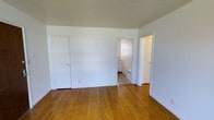 This single-floor space presents a straightforward and functional layout with a clean, understated aesthetic. The main living area is defined by warm hardwood flooring in a honey-toned finish, which runs throughout and adds a sense of natural warmth to the otherwise neutral interior. The walls are painted a crisp, bright white, contributing to an open and airy atmosphere.

Three doorways are visible from the main room: a closet to the left offers convenient built-in storage, while a central opening leads directly into what appears to be a kitchen, where tiled flooring and a radiator unit can be seen. A third door to the right provides access to an additional room. The layout connects spaces fluidly, allowing for easy movement throughout.

The ceilings are standard height, maintaining a practical and comfortable scale. The minimalist design keeps the focus on the functional elements of the space, making it well-suited for those looking to personalize it to their own taste. Overall, the space offers a clean foundation with classic hardwood floors, ample natural light, and a simple, connected floor plan.
