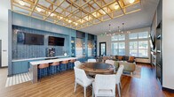 Located at 32505 Tupelo Cir in Frankford, Delaware, this single-floor community clubhouse offers a thoughtfully designed gathering space with a cohesive blend of modern and warm design elements. The main lounge area is anchored by soaring ceilings and floor-to-ceiling windows that flood the space with natural light and provide views of the outdoor pool area. A striking brick fireplace wall serves as a focal point, flanked by illuminated built-in shelving with vibrant patterned backdrops and curated décor. Seating is arranged in inviting conversation clusters across wood-look plank flooring, complemented by a sculptural multi-globe chandelier overhead. A dedicated game area features a large round gaming table with upholstered seating, positioned adjacent to a sleek wet bar finished in teal cabinetry with dark tile backsplash, bar seating, and an overhead latticed gold ceiling installation that adds architectural drama. A separate conference room provides a more intimate setting, featuring a slatted wood accent wall, wall-mounted display screen, a long communal table with blue upholstered chairs, and statement pendant lighting. Throughout, bold teal accents, warm wood tones, and layered lighting create a vibrant yet welcoming ambiance suited for both social and professional gatherings.