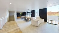 Located at 1031 N Park Ave in Tucson, Arizona, this single-floor space offers a generously sized, open-plan layout that seamlessly blends lounge and collaborative work areas. The interior is defined by a clean, contemporary aesthetic, with light hardwood flooring flowing throughout the majority of the space and a textured area rug anchoring the lounge zone.

The lounge area features plush white sofas and armchairs arranged around a brass-accented side table, creating an inviting setting for relaxed gatherings. A dramatic black-and-white typographic mural panel serves as a striking focal point, adding bold artistic character to the space. Lush tropical palms placed throughout introduce a natural, refreshing contrast against the modern architecture.

A dedicated meeting and dining area is furnished with a substantial natural wood communal table surrounded by sleek black wishbone chairs, ideal for collaborative work or group dining. Large black-framed floor-to-ceiling windows line multiple walls, flooding the interior with natural light and offering views of the surrounding Tucson streetscape.

Dark wood wall paneling provides rich contrast against the predominantly white walls and ceilings, while recessed lighting ensures a well-lit environment throughout. The space also features elevator access, making it fully accessible. Overall, the design balances minimalist sophistication with warm, functional elements, making it well-suited for professional or community-oriented use.