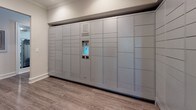 This purpose-built package locker room is designed to streamline parcel delivery and retrieval for building residents or occupants. The centerpiece of the space is an expansive Package Concierge automated locker system, which spans nearly the full length of multiple walls, offering a generous array of secure, individually sized compartments in varying heights to accommodate packages of different dimensions. The system includes an integrated touchscreen kiosk, allowing users to conveniently retrieve their deliveries at any time. The lockers feature a clean, modern aesthetic with a uniform light gray finish that complements the room's neutral color palette of soft whites and warm beige tones. The flooring throughout is a durable wood-look plank material in a gray-toned finish, lending a polished yet practical feel to the space. Crown molding and white baseboards add a refined architectural touch, maintaining consistency with the building's overall interior design. Security is reinforced by a ceiling-mounted surveillance camera. The room connects directly to adjacent common areas, including what appears to be a lounge or sitting area, ensuring seamless integration into the building's overall layout. The space is well-lit with recessed overhead lighting, creating a bright and accessible environment.