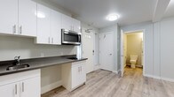 This thoughtfully designed single-floor space offers a clean, contemporary aesthetic with a bright and functional layout. Light hardwood-style flooring runs consistently throughout, tying each area together with a cohesive, modern feel.

The kitchenette is a standout feature, equipped with crisp white cabinetry extending from floor to ceiling, providing generous storage. Dark charcoal countertops offer a striking contrast to the white cabinetry, while a stainless steel sink, built-in cooktop, and over-the-range microwave deliver practical, efficient functionality. The entry door beside the kitchenette features a transom window, allowing natural light to filter into the space.

The open living area is versatile and unencumbered, with neutral light gray walls and clean white baseboards creating a calm, adaptable backdrop. Flush-mount ceiling light fixtures provide consistent illumination throughout. The space's open configuration allows for flexible furniture arrangement to suit a variety of uses.

The bathroom maintains the same neutral palette, with light gray walls and matching hardwood-style flooring. It features a wall-mounted grab bar for added accessibility and convenience, along with a standard toilet. A bifold door separates the bathroom from the rest of the unit, maximizing space efficiency. Overall, the unit presents a well-appointed, move-in-ready environment with modern finishes and practical design choices throughout.