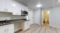 This thoughtfully designed single-floor space offers a clean, contemporary aesthetic with a bright and functional layout. Light hardwood-style flooring runs consistently throughout, tying each area together with a cohesive, modern feel.

The kitchenette is a standout feature, equipped with crisp white cabinetry extending from floor to ceiling, providing generous storage. Dark charcoal countertops offer a striking contrast to the white cabinetry, while a stainless steel sink, built-in cooktop, and over-the-range microwave deliver practical, efficient functionality. The entry door beside the kitchenette features a transom window, allowing natural light to filter into the space.

The open living area is versatile and unencumbered, with neutral light gray walls and clean white baseboards creating a calm, adaptable backdrop. Flush-mount ceiling light fixtures provide consistent illumination throughout. The space's open configuration allows for flexible furniture arrangement to suit a variety of uses.

The bathroom maintains the same neutral palette, with light gray walls and matching hardwood-style flooring. It features a wall-mounted grab bar for added accessibility and convenience, along with a standard toilet. A bifold door separates the bathroom from the rest of the unit, maximizing space efficiency. Overall, the unit presents a well-appointed, move-in-ready environment with modern finishes and practical design choices throughout.