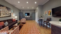 This Nashville-themed media room offers a welcoming, well-appointed communal space designed for both relaxation and social gathering. The room is finished with warm hardwood-style flooring, neutral gray walls, and crown molding, creating a polished yet comfortable atmosphere throughout.

The main lounge area features a plush dark sofa and patterned accent chairs arranged around a wood coffee table, all anchored by a bold orange geometric area rug. A large photographic print of the iconic Ryman Auditorium serves as a focal point on the wall, accompanied by an eclectic collection of music concert and Nashville travel posters that reinforce the room's distinct musical identity.

Along one wall, built-in gray cabinetry with butcher block countertops flanks a wall-mounted flat-screen television, providing ample storage and a dedicated media viewing area. Recessed lighting illuminates the space evenly from above.

A high-top bar ledge with red leather stools lines one wall beneath a trio of framed Nashville and Music City vintage-style posters, offering a casual perch for conversation or viewing. Additional high-top tables with leather chairs are positioned nearby, adding flexible seating options. A door with glass panels provides access to an outdoor area, allowing natural light to filter into the room. The overall ambiance is lively, curated, and distinctly Nashville in character.